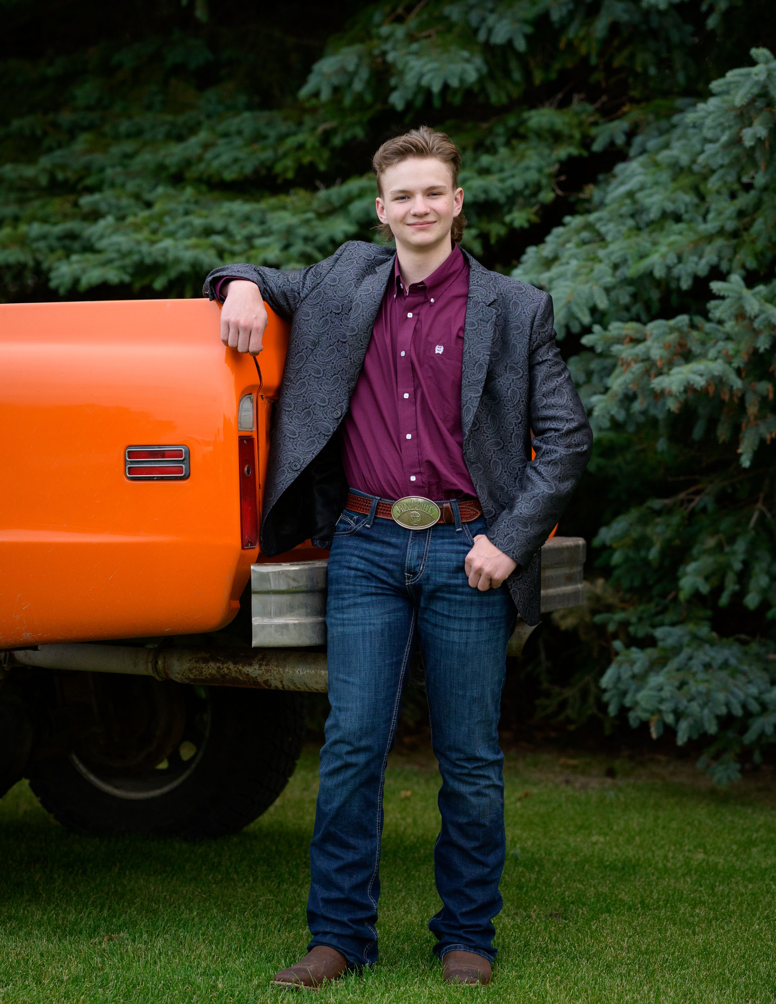 A figure poses next to an orange tractor wearing a grey blazer and burgundy shirt.
