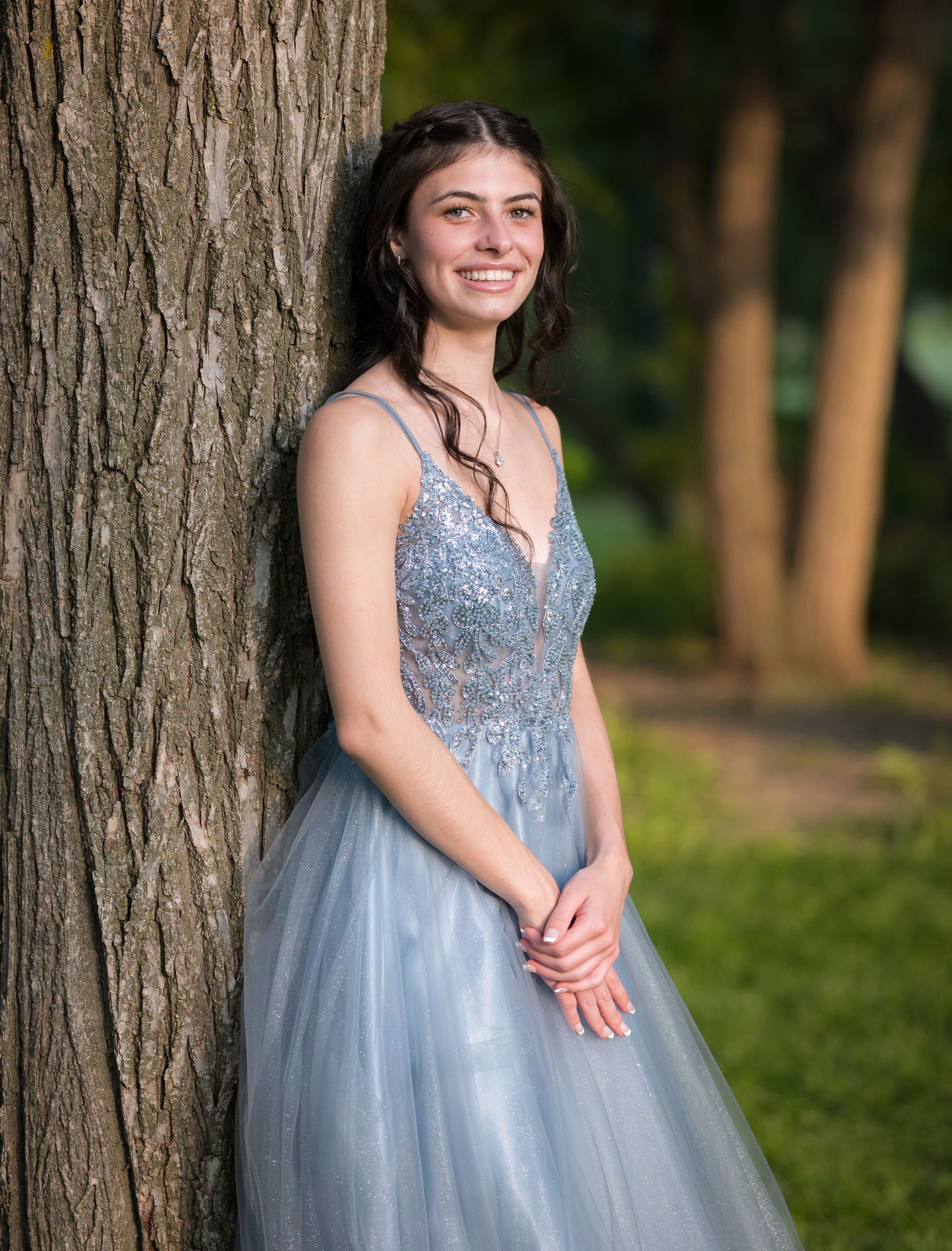 A person in a sparkly light blue formal gown stands by a large tree trunk in an outdoor setting.
