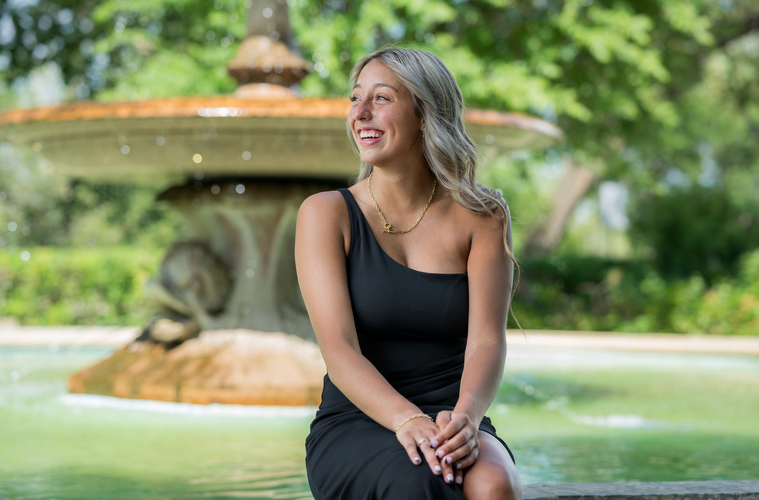 A young woman in a black dress sits outdoors by a garden fountain on a sunny afternoon.
