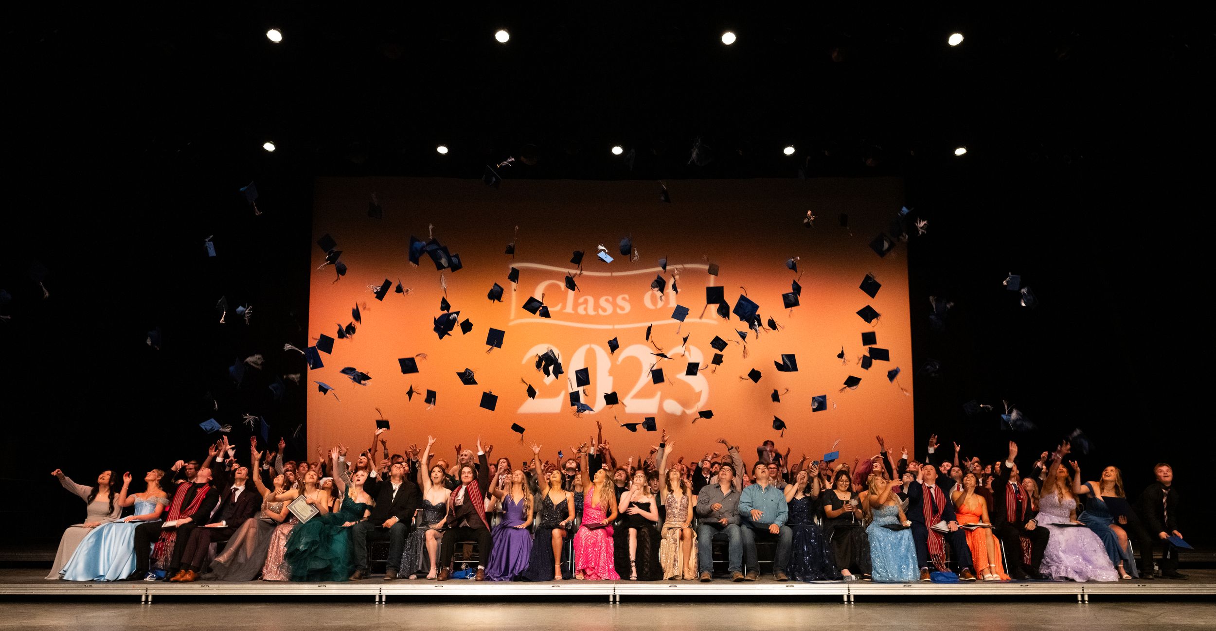 A large group of graduates celebrate by throwing their caps in the air against an orange-lit backdrop on stage.