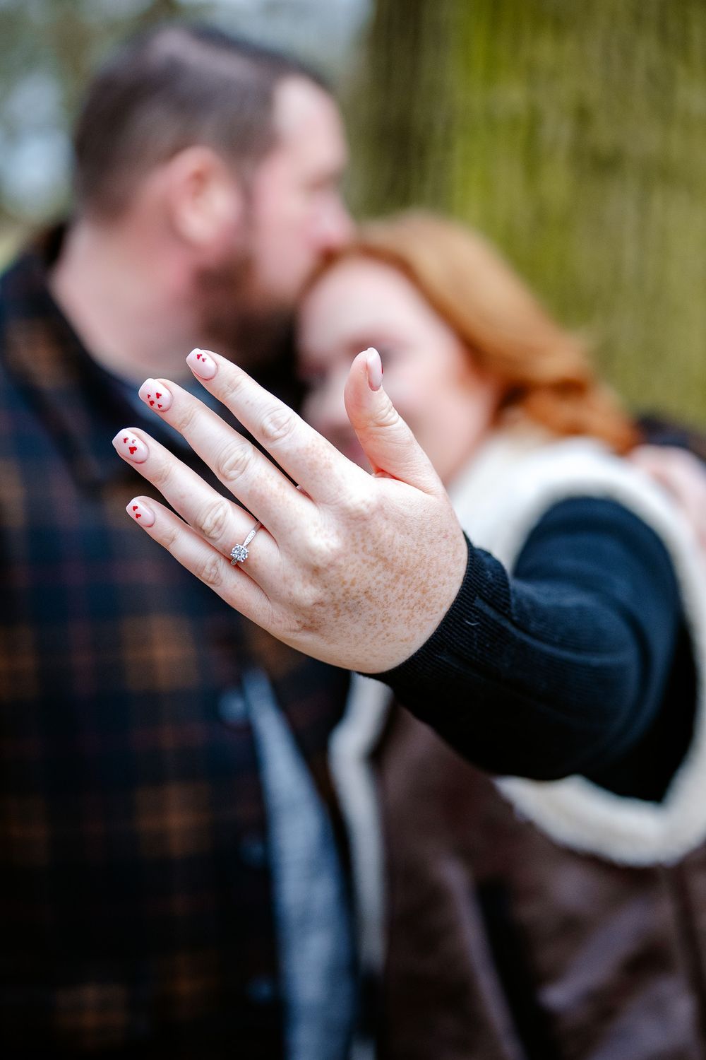 Close-up of a hand displaying an engagement ring with decorated nails against a blurred background.