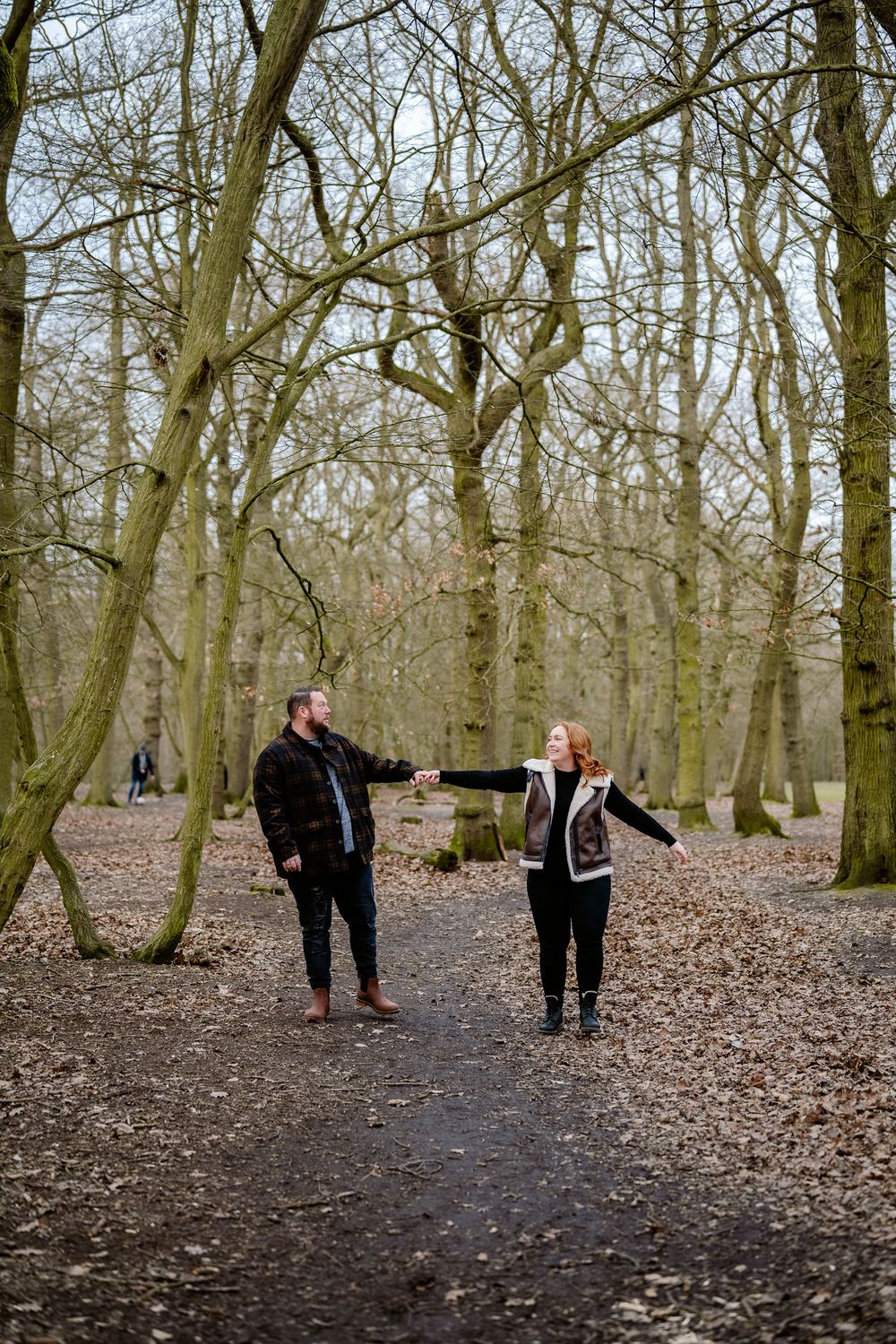 Two people playfully pose with outstretched arms on a forest path covered in fallen leaves.