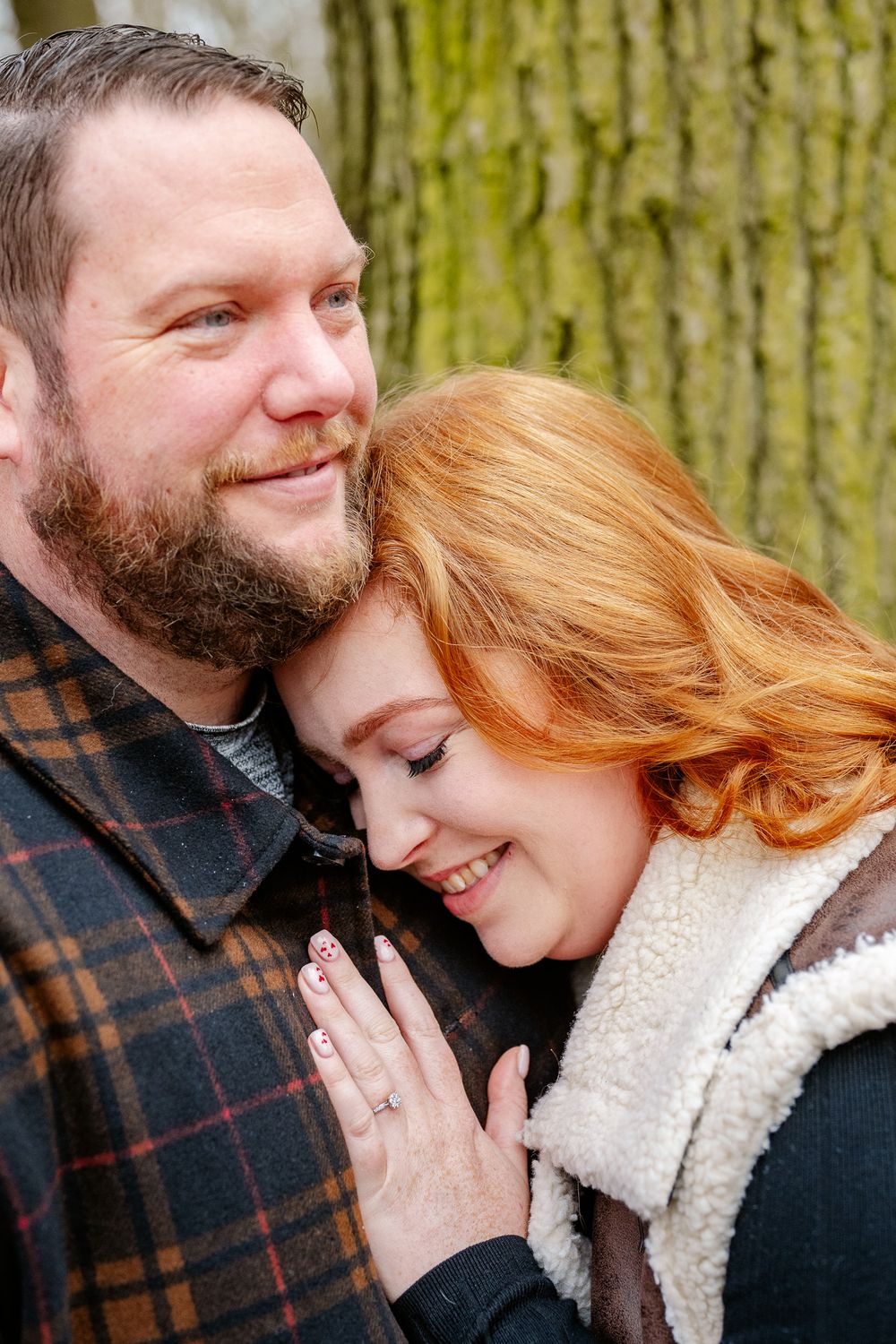 A couple in winter clothing share an intimate moment while embracing near a moss-covered tree.