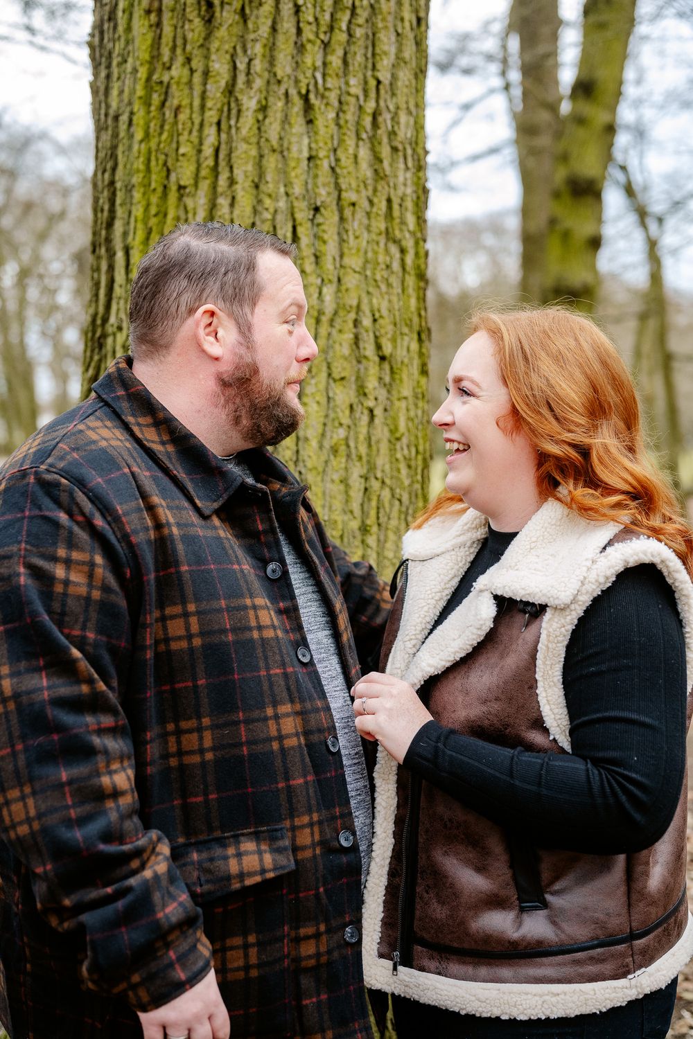 A couple in cozy winter attire share a joyful moment while standing against a mossy tree trunk in the woods.