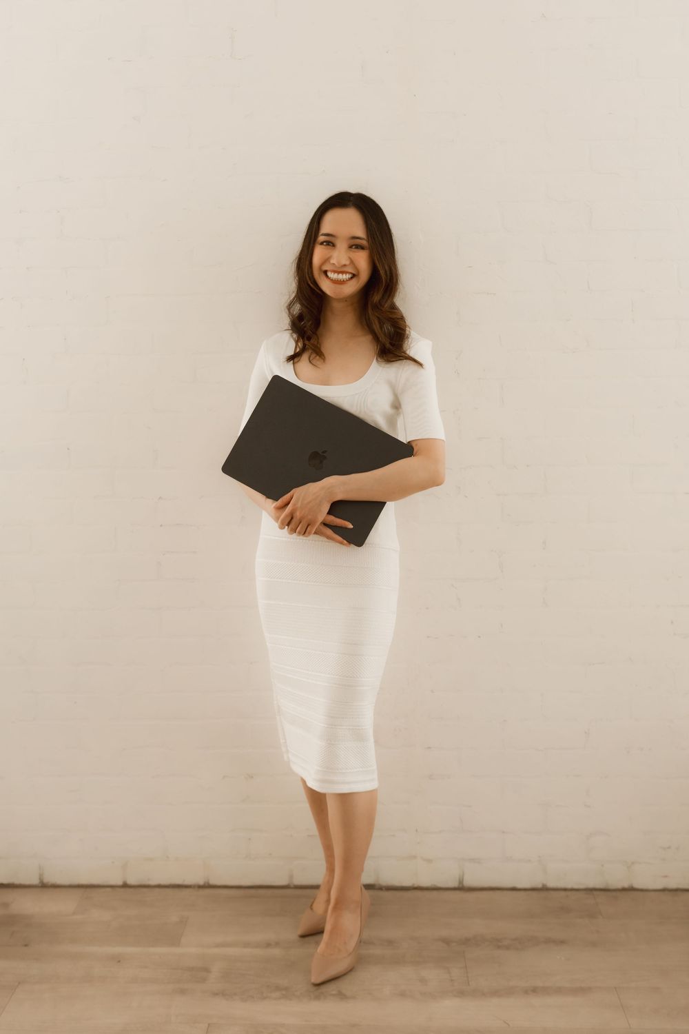 Professional in a white midi dress holding a dark portfolio folder stands against a neutral wall with beige heels.