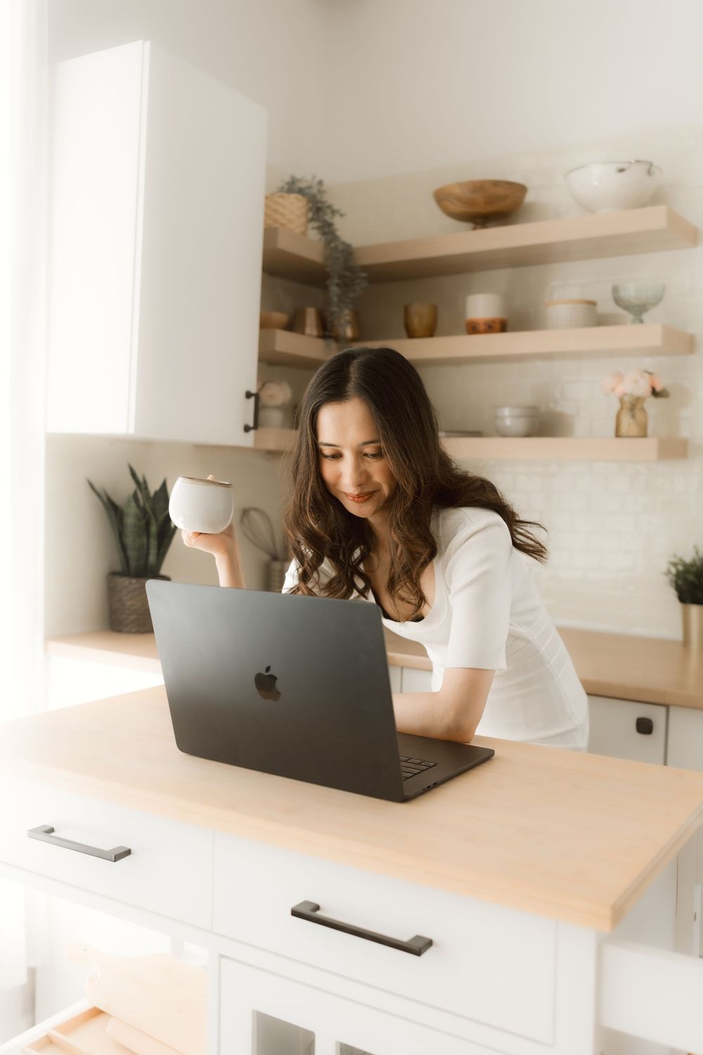 Remote worker using MacBook laptop while drinking coffee in bright modern kitchen with floating shelves.