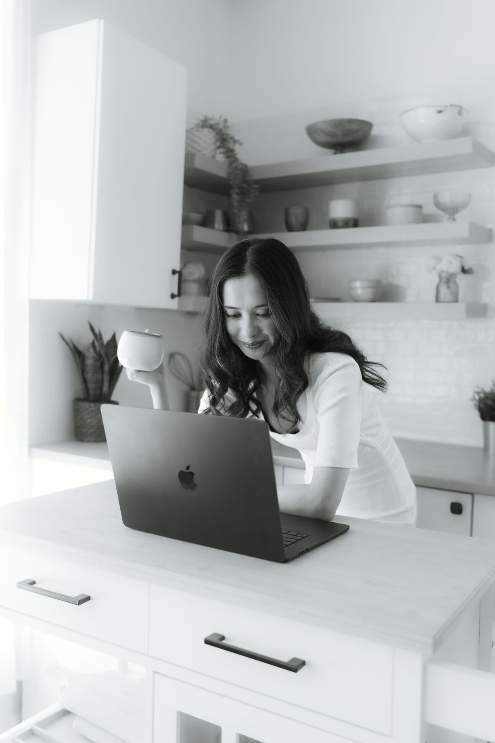 Black and white photo of person working on MacBook in modern kitchen with floating shelves.
