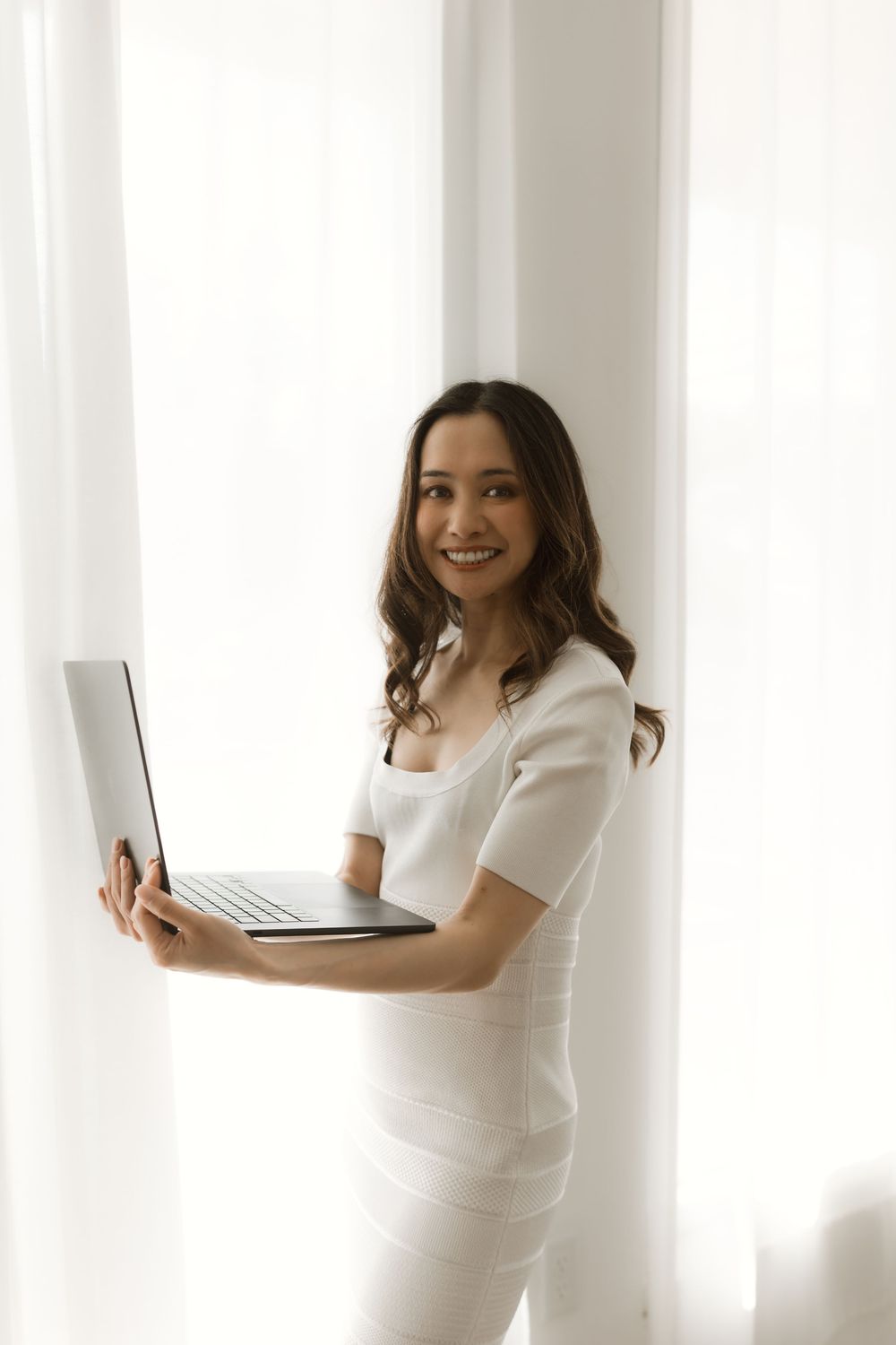 Professional in white outfit smiling while holding laptop computer near bright window.