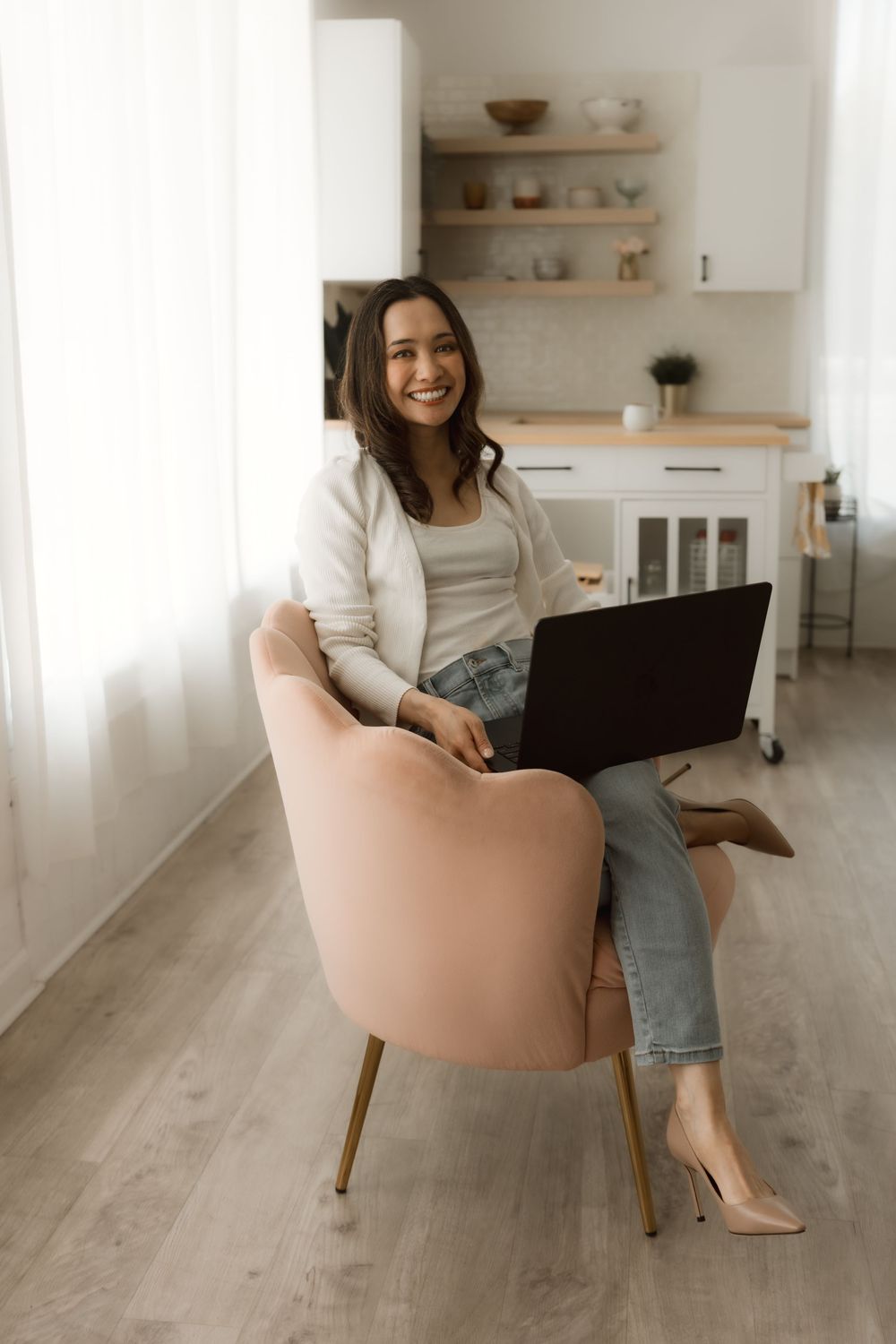 Remote worker in casual attire sitting in peach accent chair with laptop in minimalist home office.