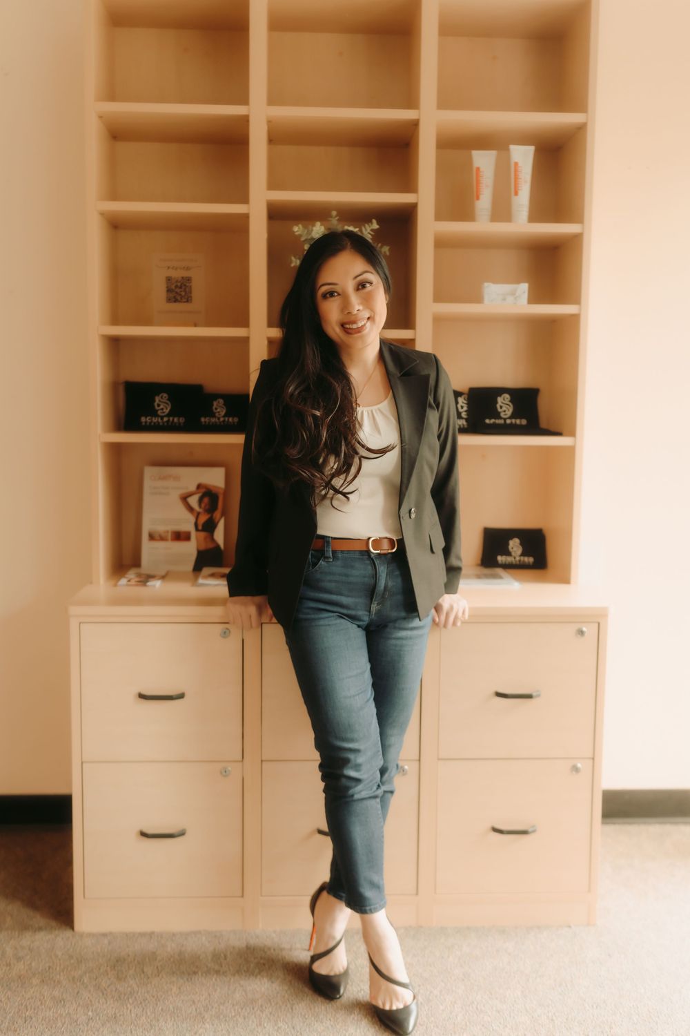 Professional lifestyle portrait in front of built-in shelving wearing blazer, jeans and white top in warm lighting.