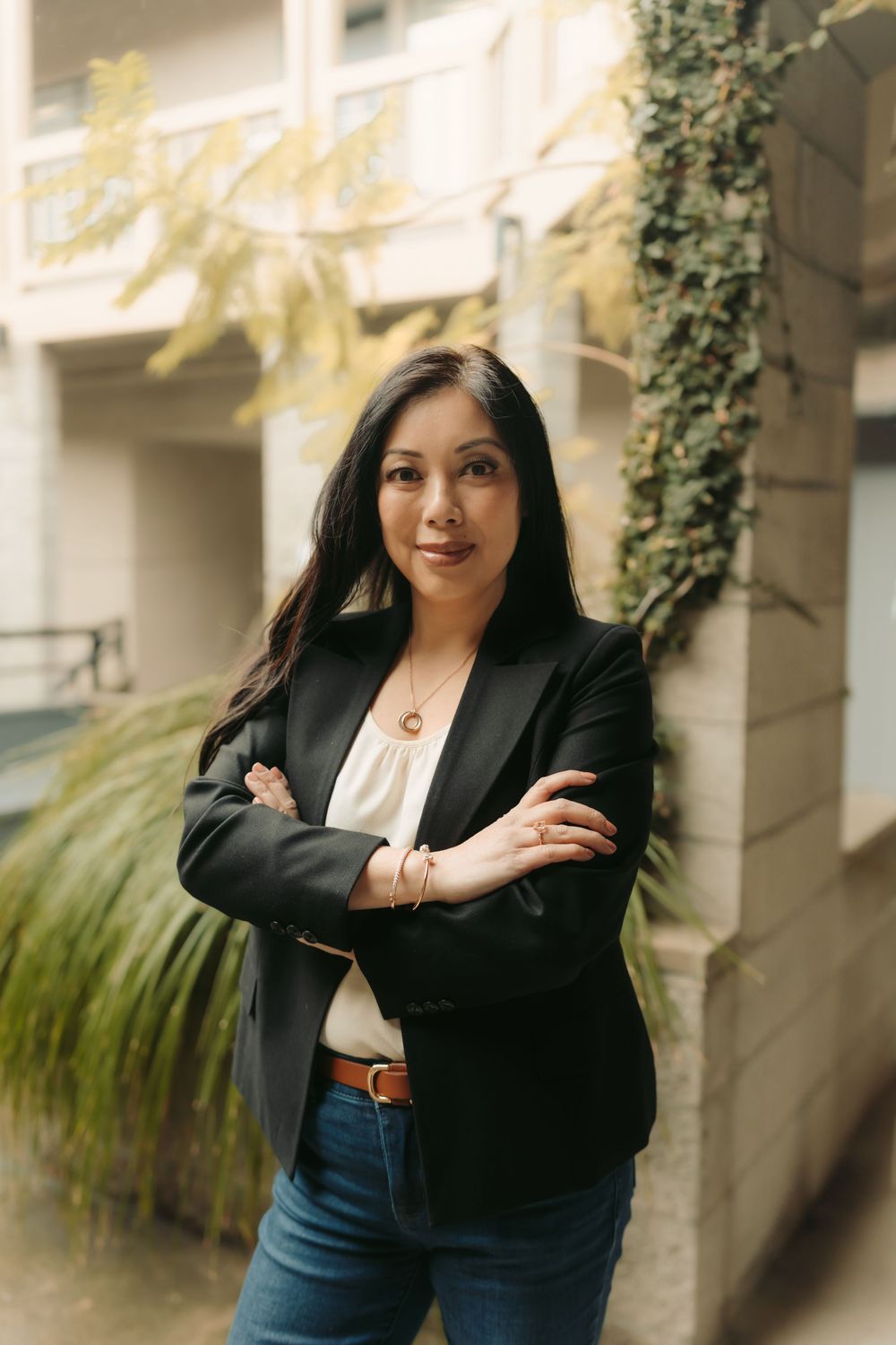 Professional woman in black blazer and jeans stands confidently against ivy-covered wall outdoors.