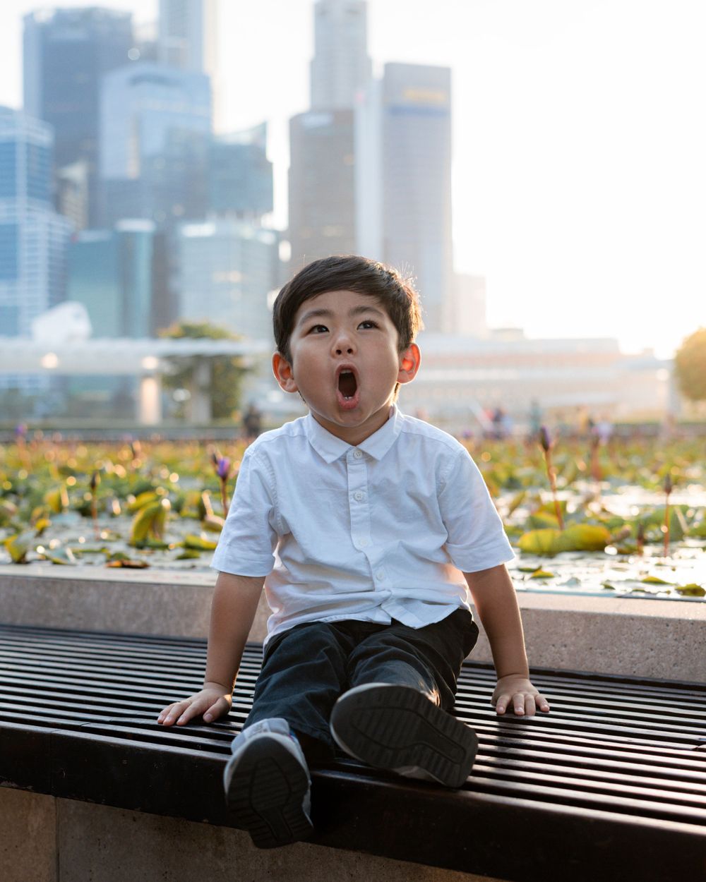 Child sitting on bench making excited expression with city skyline in background.