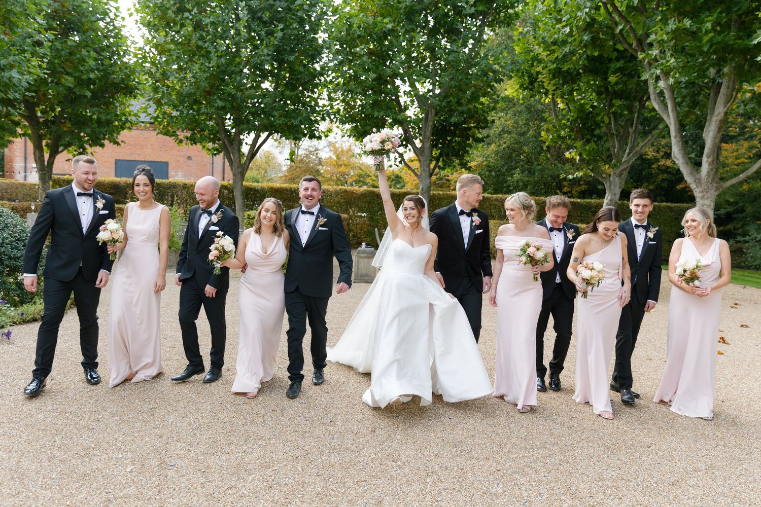 Wedding party in formal attire poses together outdoors on a gravel path with trees in the background.