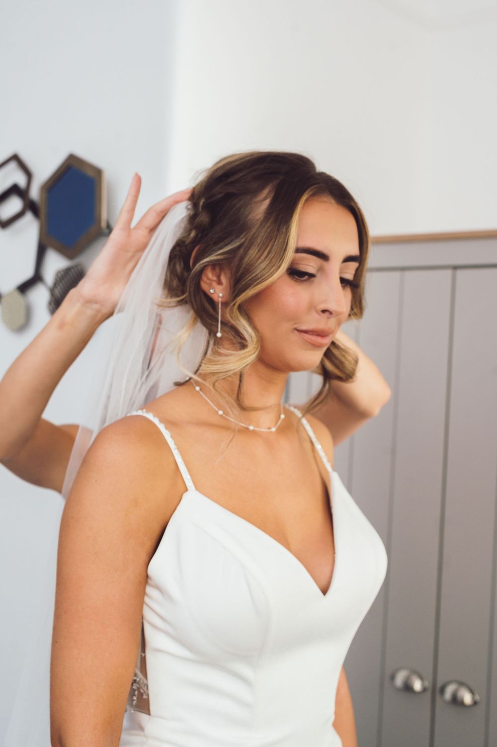 A bride in a white spaghetti strap wedding dress and veil adjusts her hair while wearing a delicate necklace.