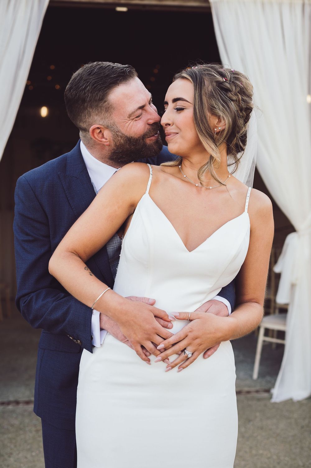 Romantic wedding couple embraces in front of white curtains, bride wearing a sleek white gown.