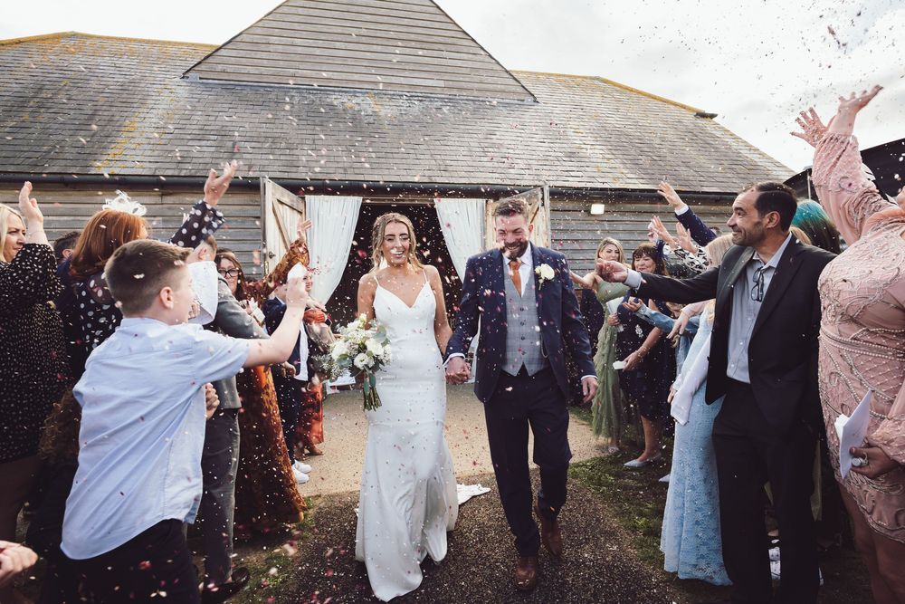 Wedding guests throw confetti over newlyweds as they exit a rustic barn venue on their special day.