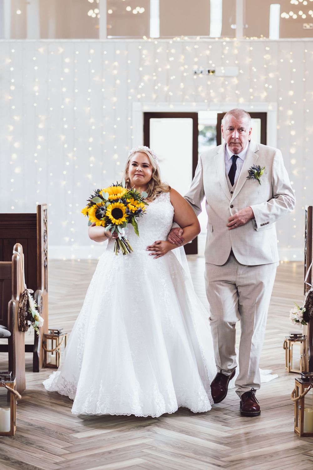 A wedding couple walks down a wooden floor aisle with sunflowers and twinkle lights in the background.