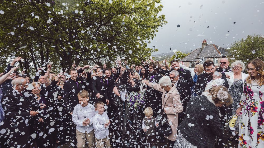 A large group of wedding guests celebrate outdoors as confetti rains down during a joyous celebration.