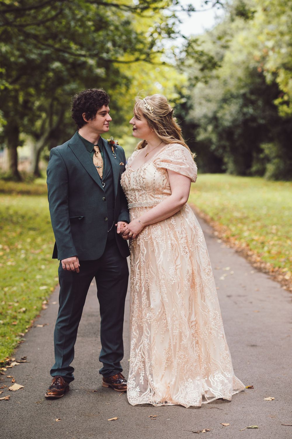 A couple in formal attire walks along a tree-lined path with autumn leaves scattered on the ground.