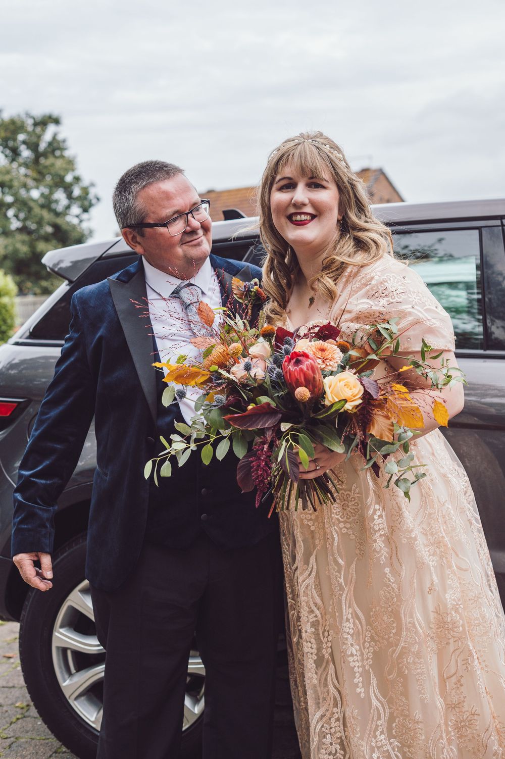 A couple shares a joyful moment outside their car on their wedding day, with the bride holding a vibrant autumn bouquet.