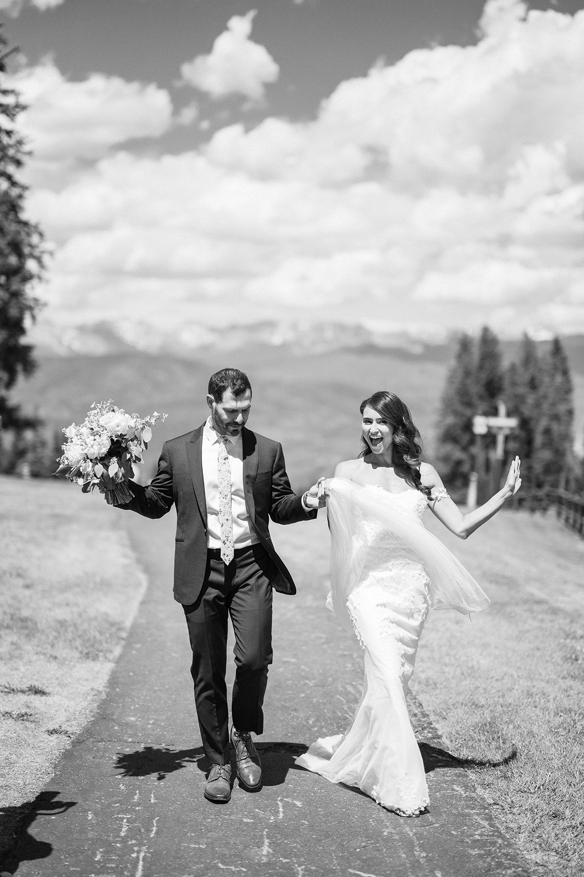 A celebratory wedding couple dances on a mountain path with scenic views.