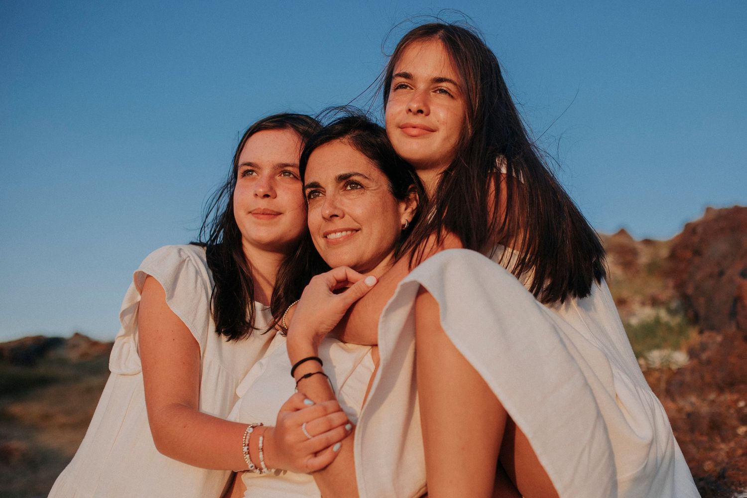 Three people in white outfits pose together against a blue sky and desert landscape during golden hour.