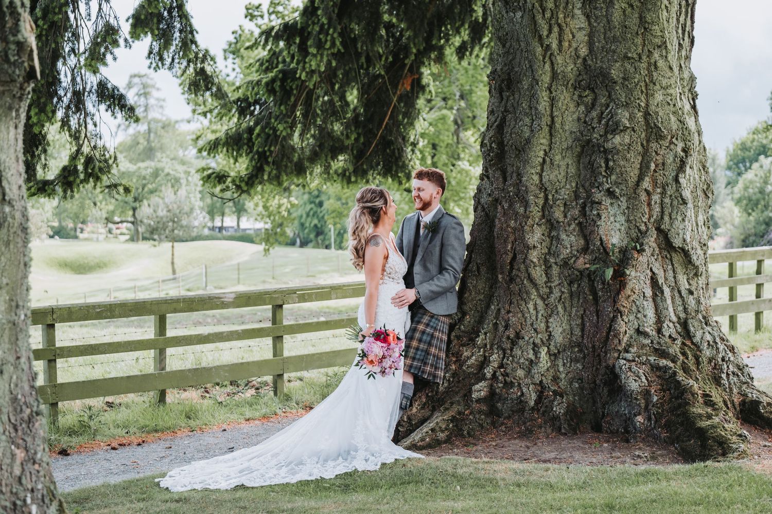 Bride and groom standing by a large tree in a lush garden, bride holding a red bouquet in a white gown.