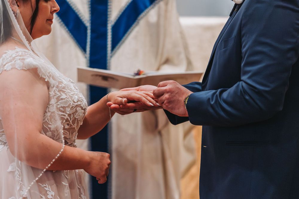 Close-up view of hands exchanging wedding rings during a marriage ceremony with lace bridal gown and navy suit details.