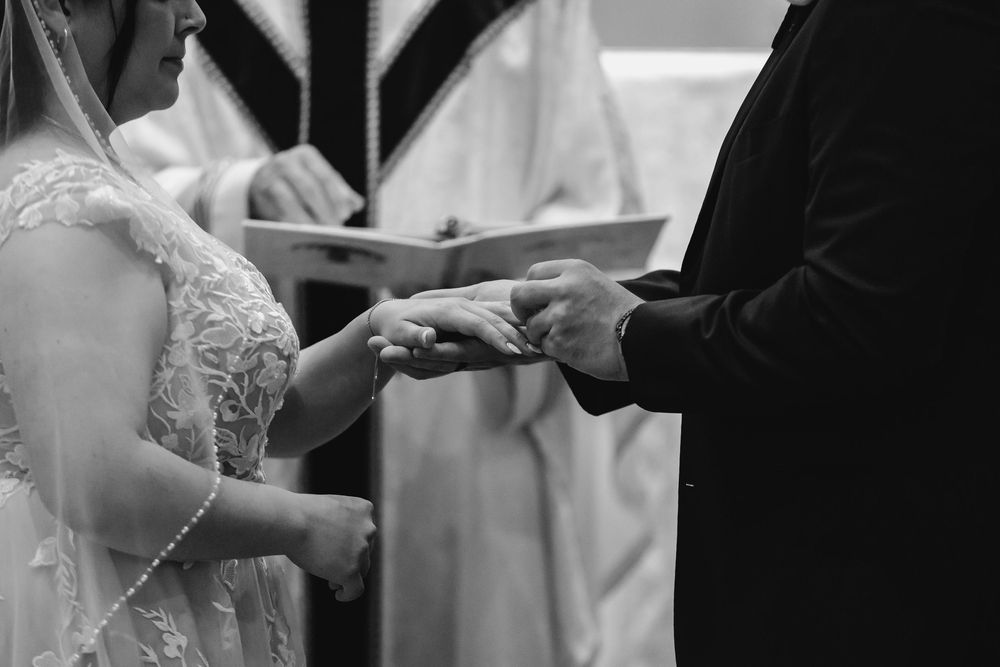 Two people exchange rings during a wedding ceremony while dressed in formal white attire.