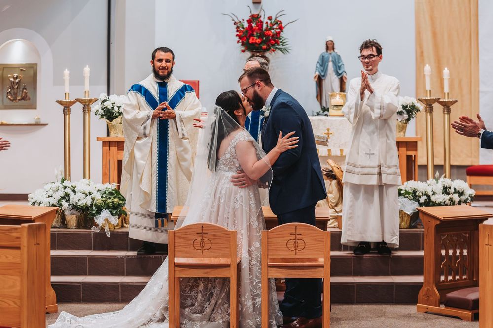A couple shares their first kiss during a Catholic wedding ceremony inside a church decorated with flowers and candles.
