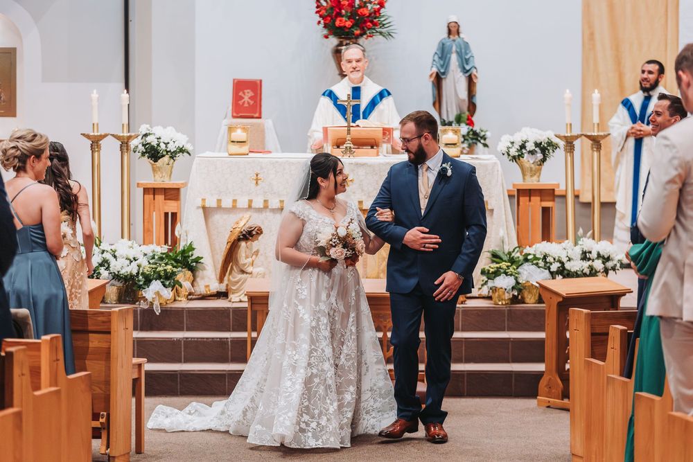 A couple stands together during their wedding ceremony in a church decorated with white floral arrangements and religious statues.