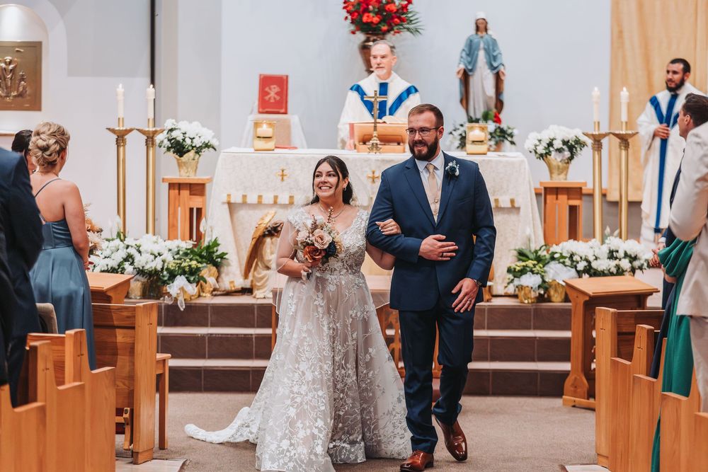 A couple walks down the church aisle during a wedding ceremony with floral arrangements decorating the altar.