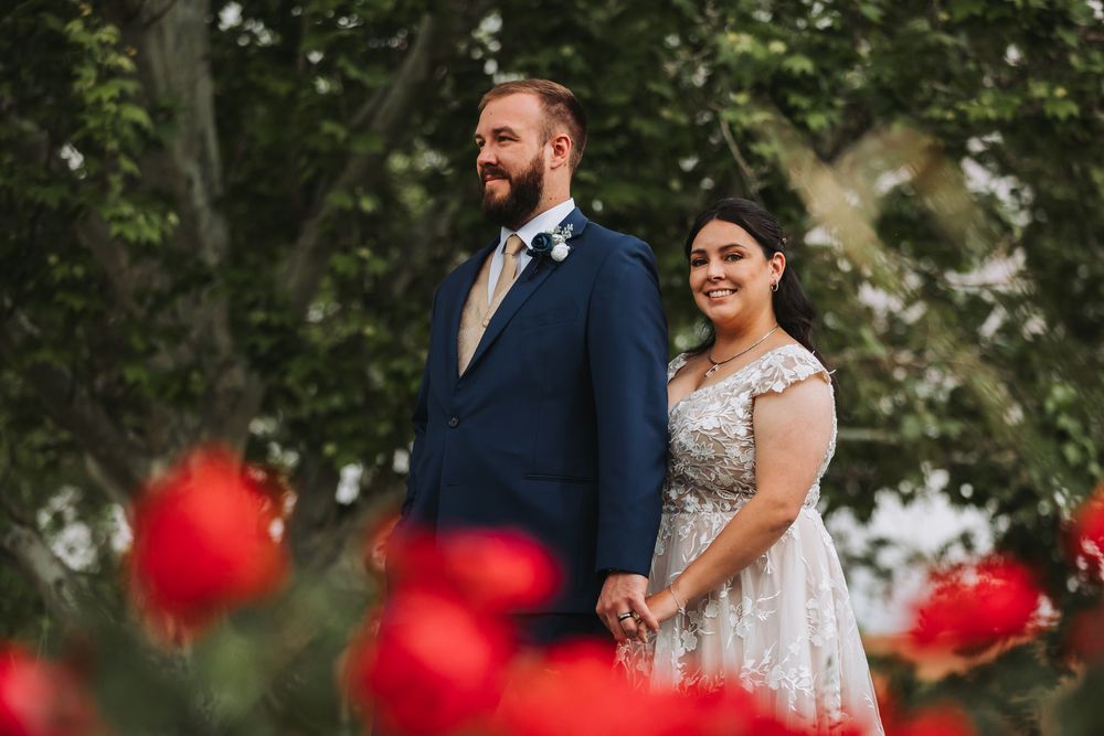 A bride and groom hold hands while standing in a garden with red flowers in the foreground.