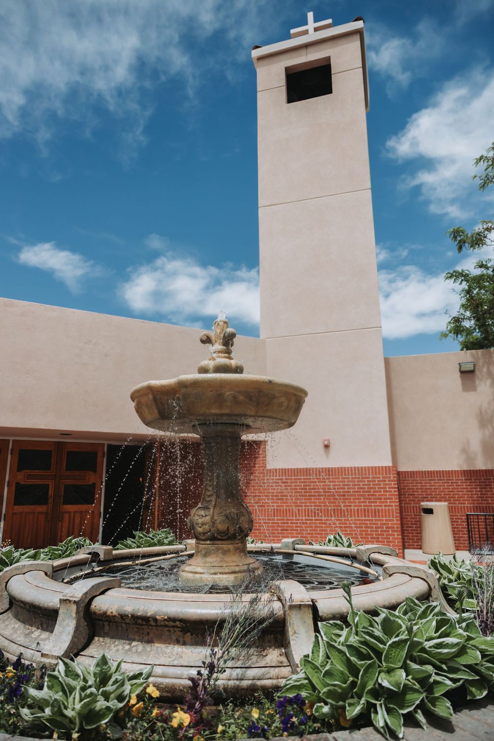 White church steeple rises above a decorative stone fountain surrounded by lush hostas and flowers under blue skies.
