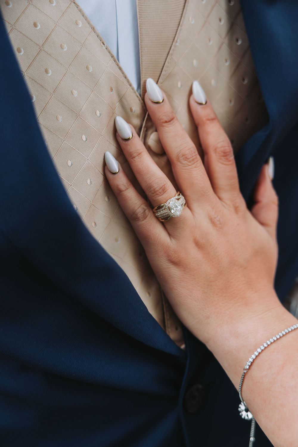 A manicured hand with white nail polish and diamond ring rests against a navy and beige textured fabric background.