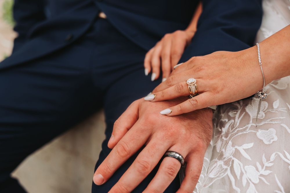 Closeup of hands displaying wedding rings against a white lace dress background.