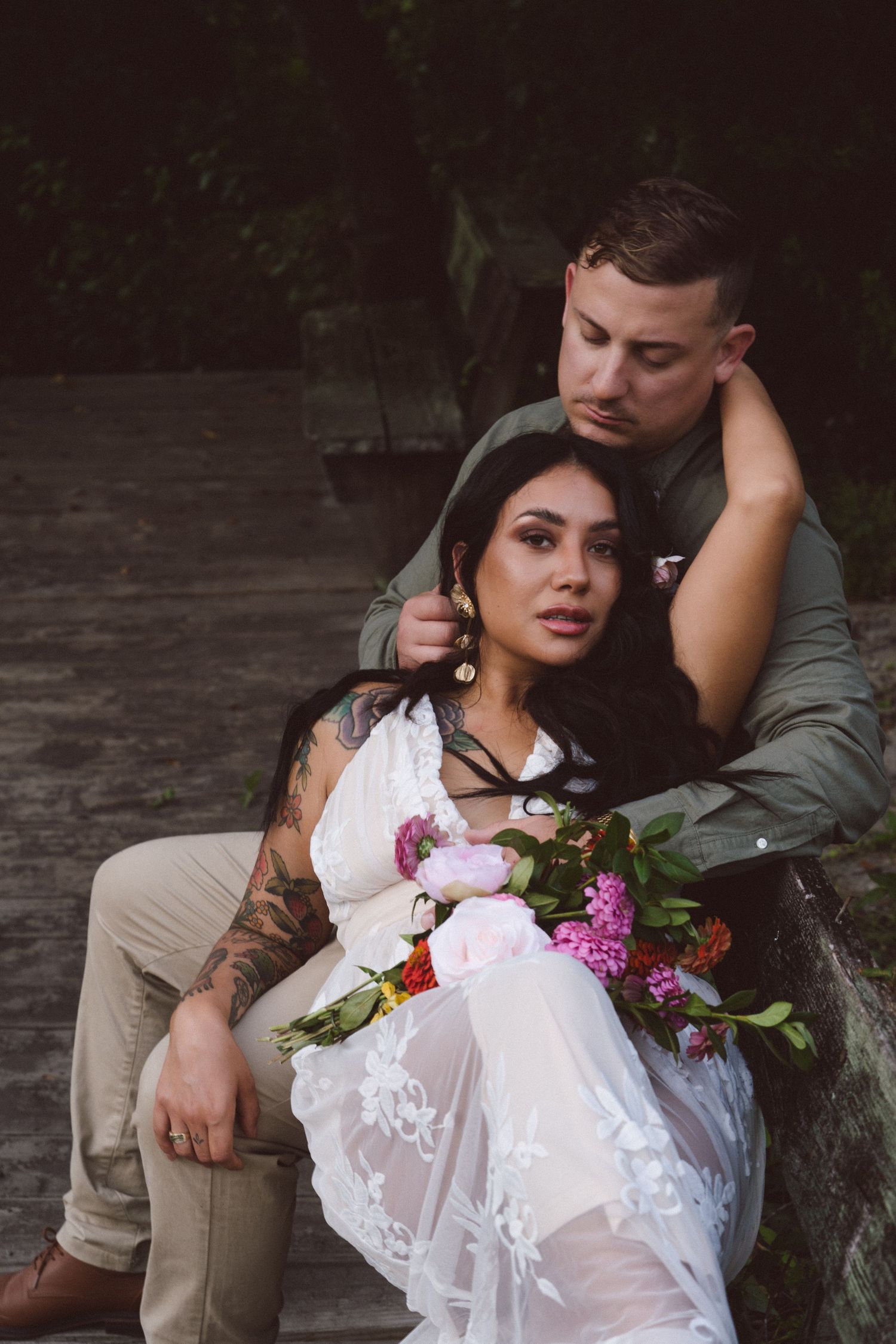 A romantic couple sits by a waterside rock, the woman in white lace holding pink and purple flowers in her lap.
