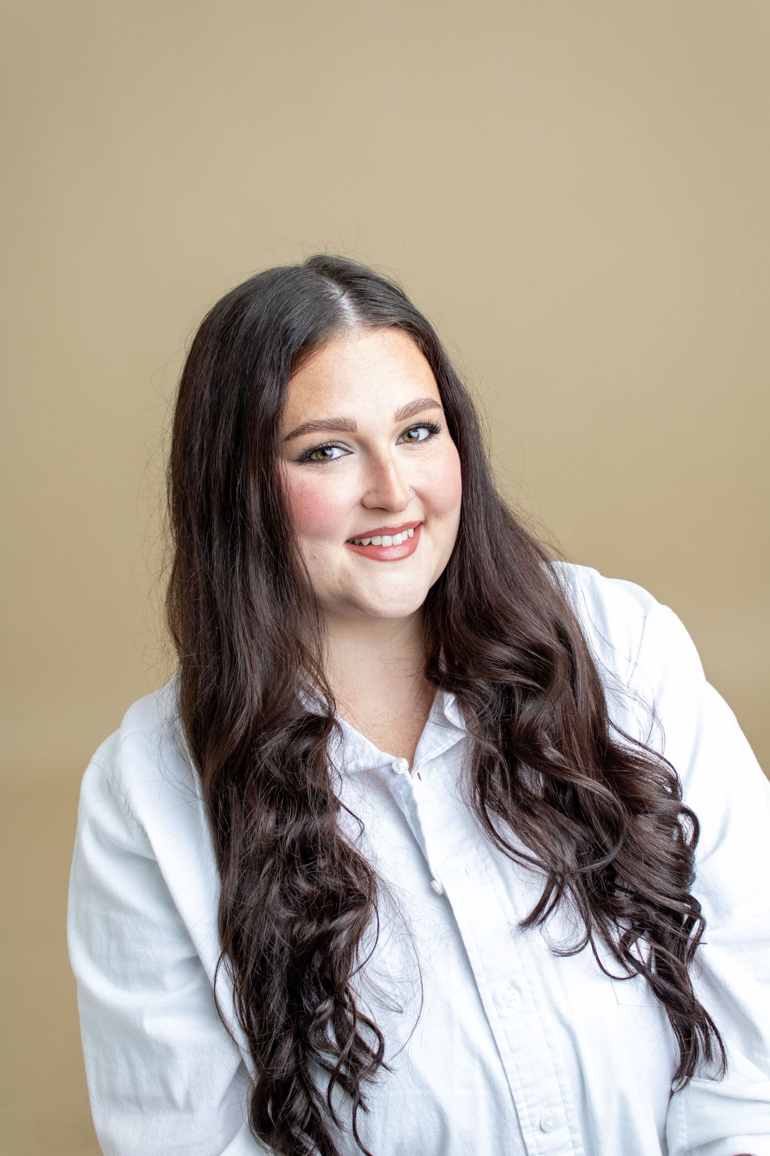 A young woman with long dark wavy hair smiles warmly wearing a white button-up shirt on a beige background.