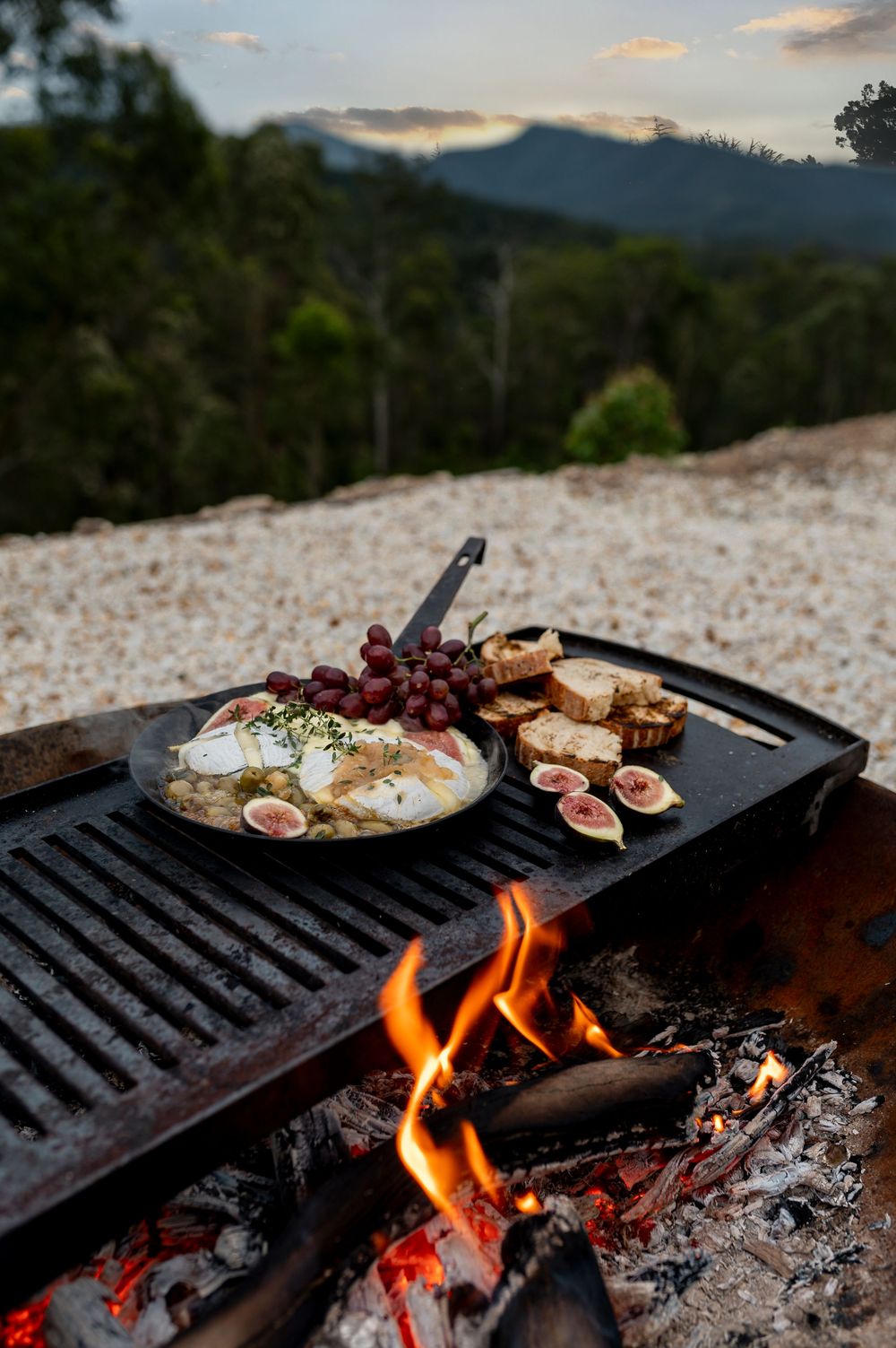 Grilled brie cheese with grapes and figs cooking over an open campfire with mountain views at sunset.