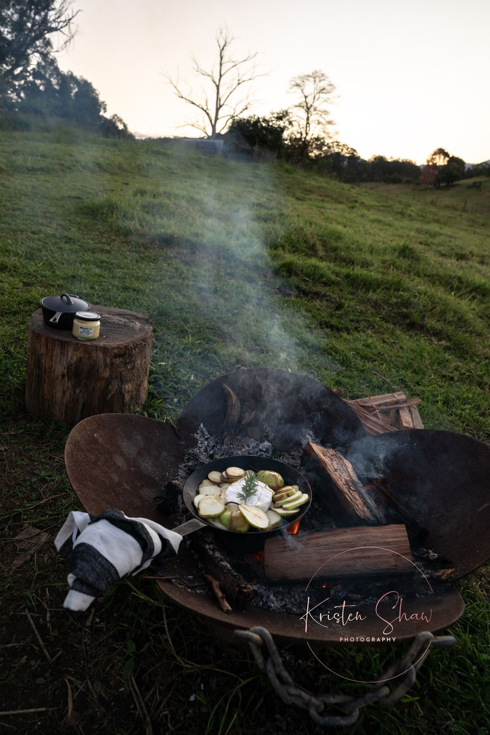 Fresh ingredients cooking over a rustic campfire with smoke rising on a grassy hillside at sunset.
