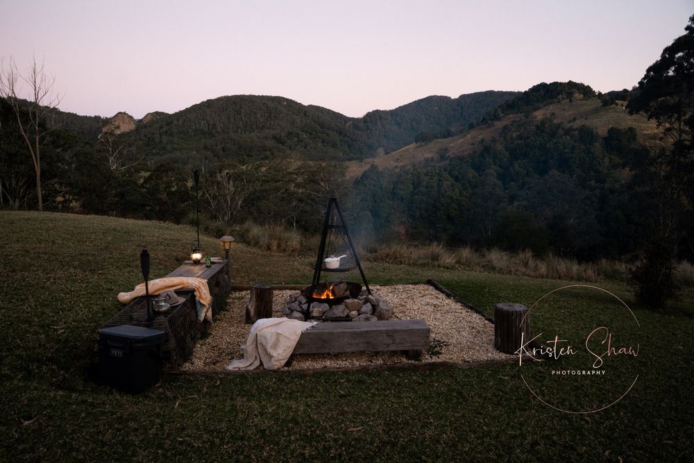 Rustic outdoor campfire setup with cooking pot on tripod against mountain landscape at dusk.