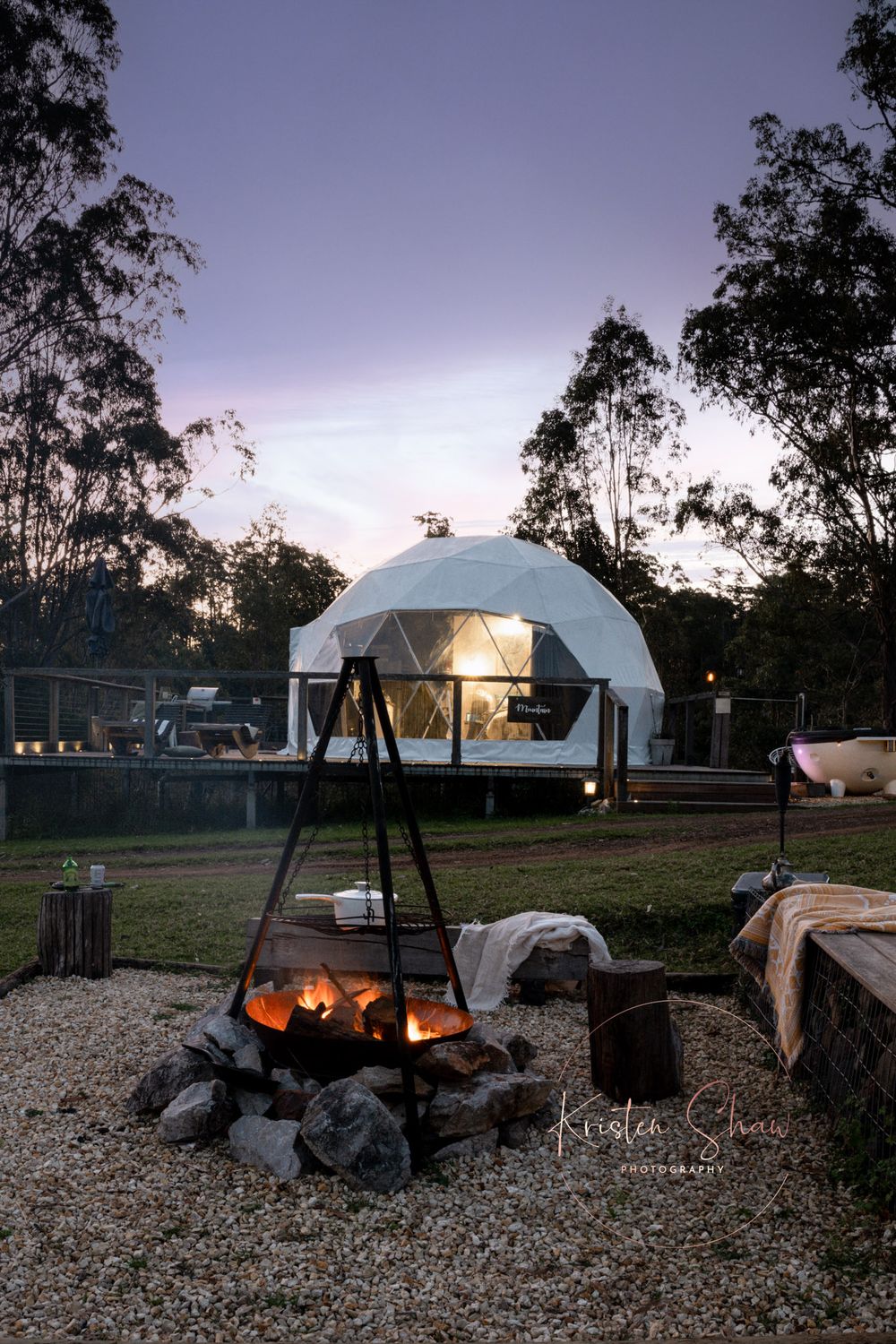 A campfire burns in front of a geodesic dome tent at dusk, with eucalyptus trees silhouetted against the purple sky.