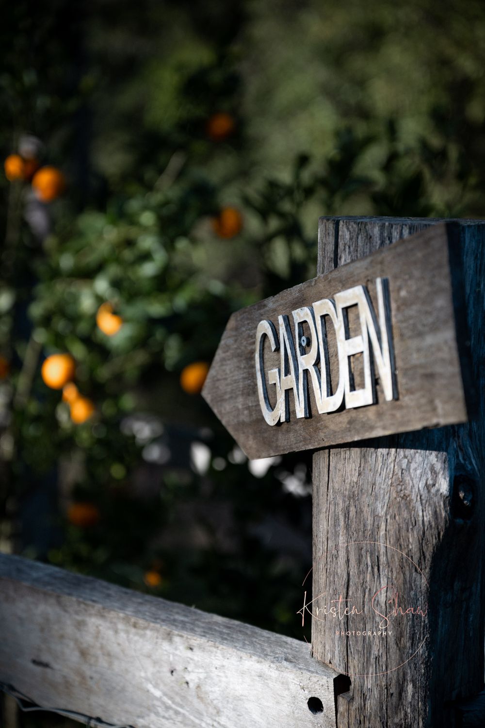 Rustic wooden garden sign with orange citrus fruits blurred in the background.
