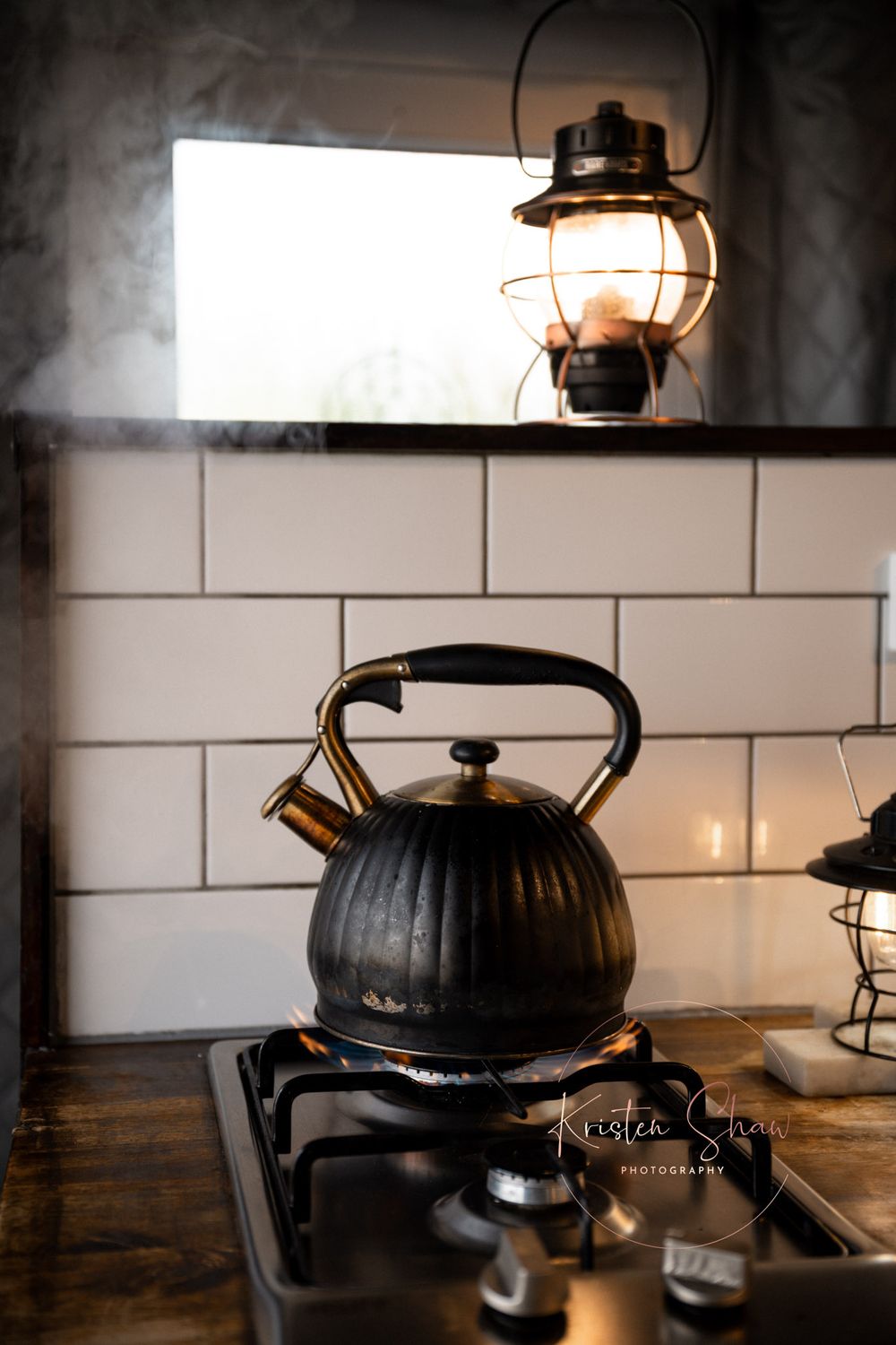 Black cast iron teapot with a vintage lantern light fixture on dark wood surface in rustic kitchen setting.