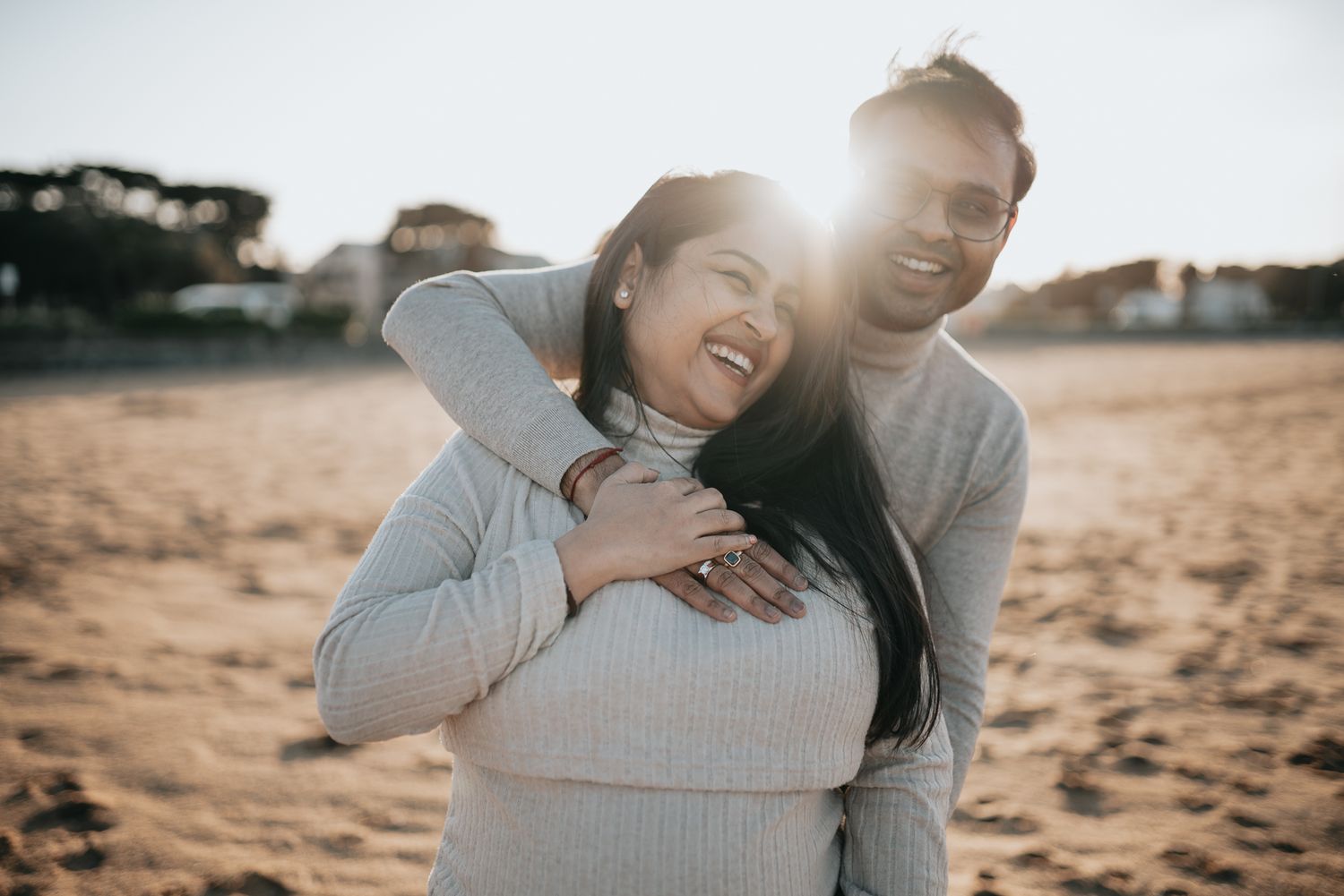 Young couple laughing together on sandy beach at sunset wearing matching gray sweaters taken by Melbourne maternity photographer.