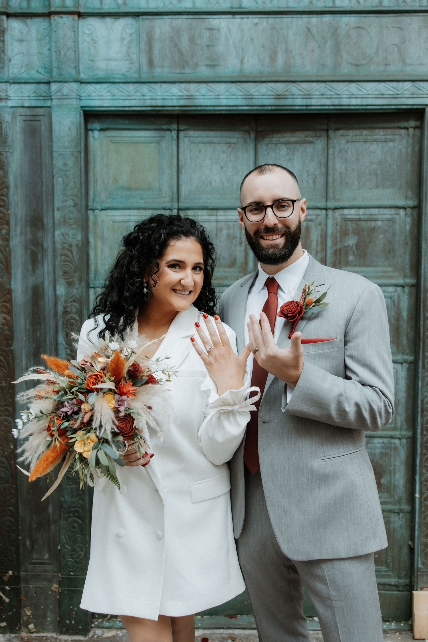 Newlywed couple in white dress and gray suit showing off wedding rings against teal wall.