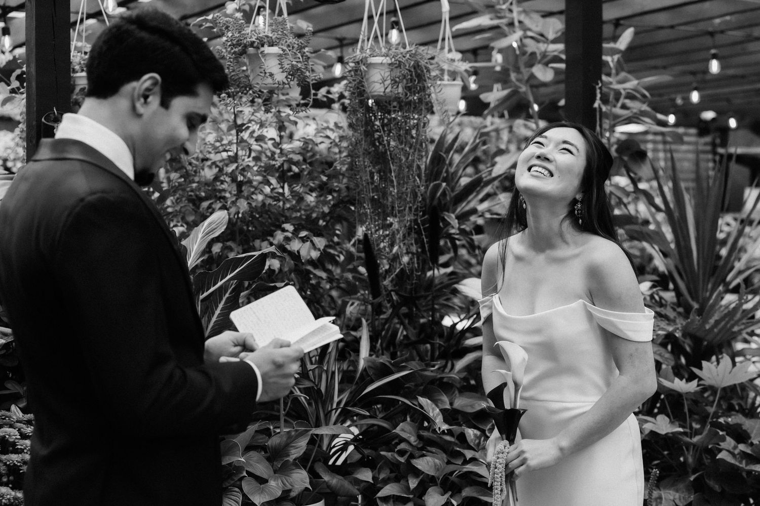 A black and white wedding moment captured in a greenhouse setting with lush plants and flowers surrounding the couple.