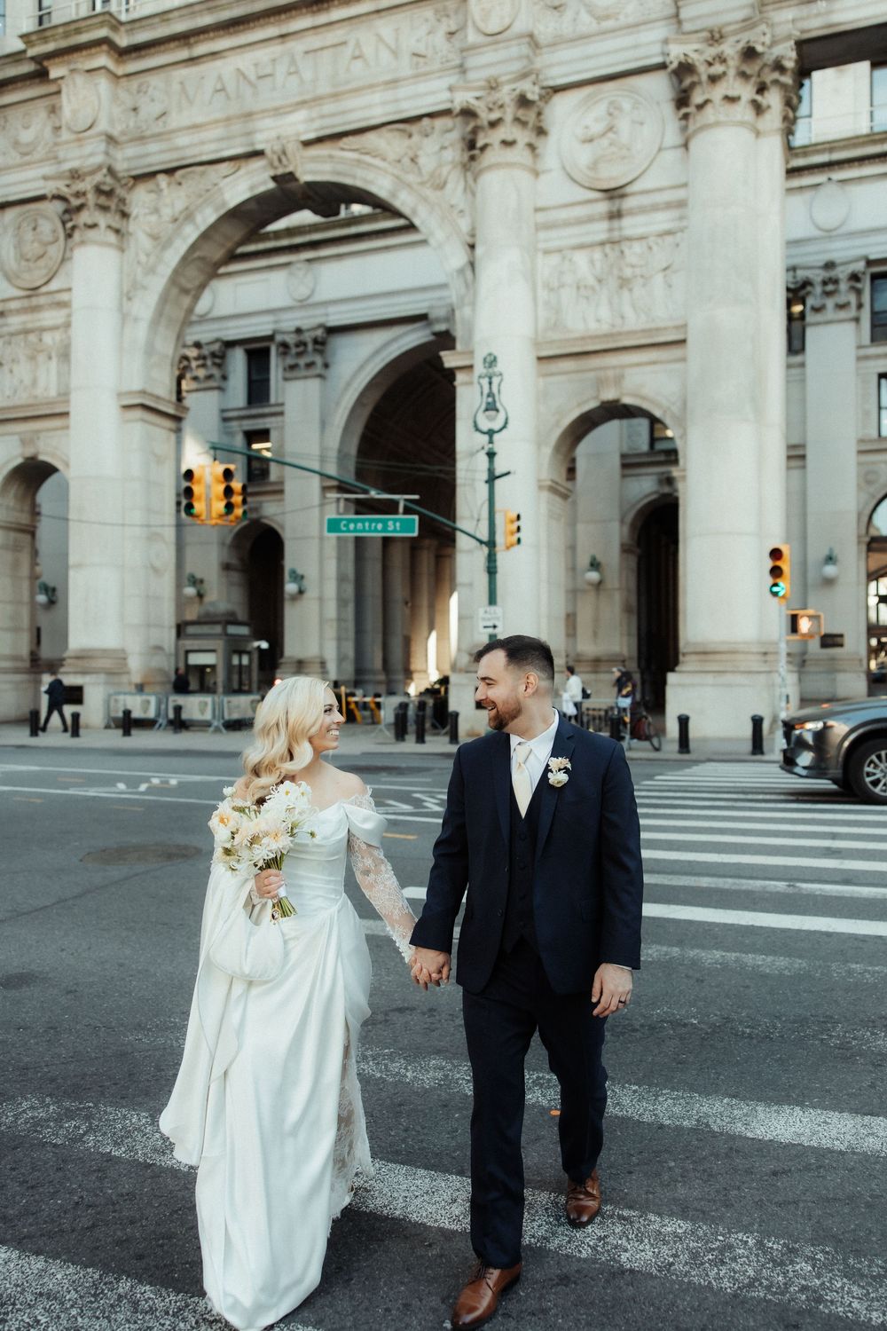 A couple in wedding attire holds hands while crossing a city street near an ornate stone archway with traffic lights.