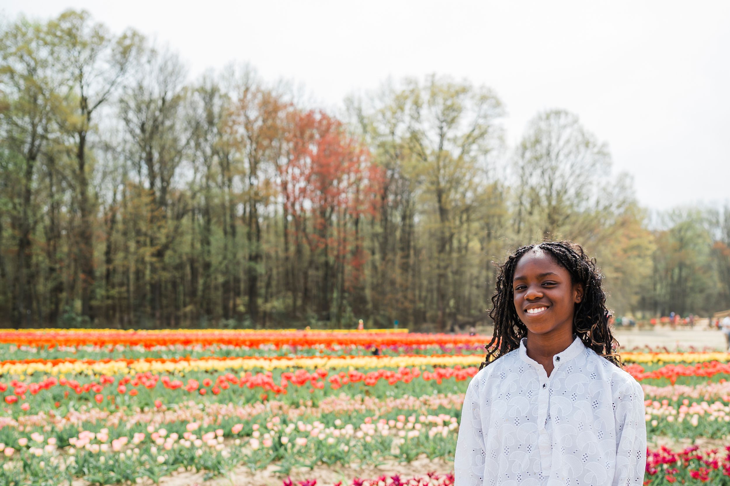 Person in white shirt standing in front of colorful tulip field with fall foliage in background.