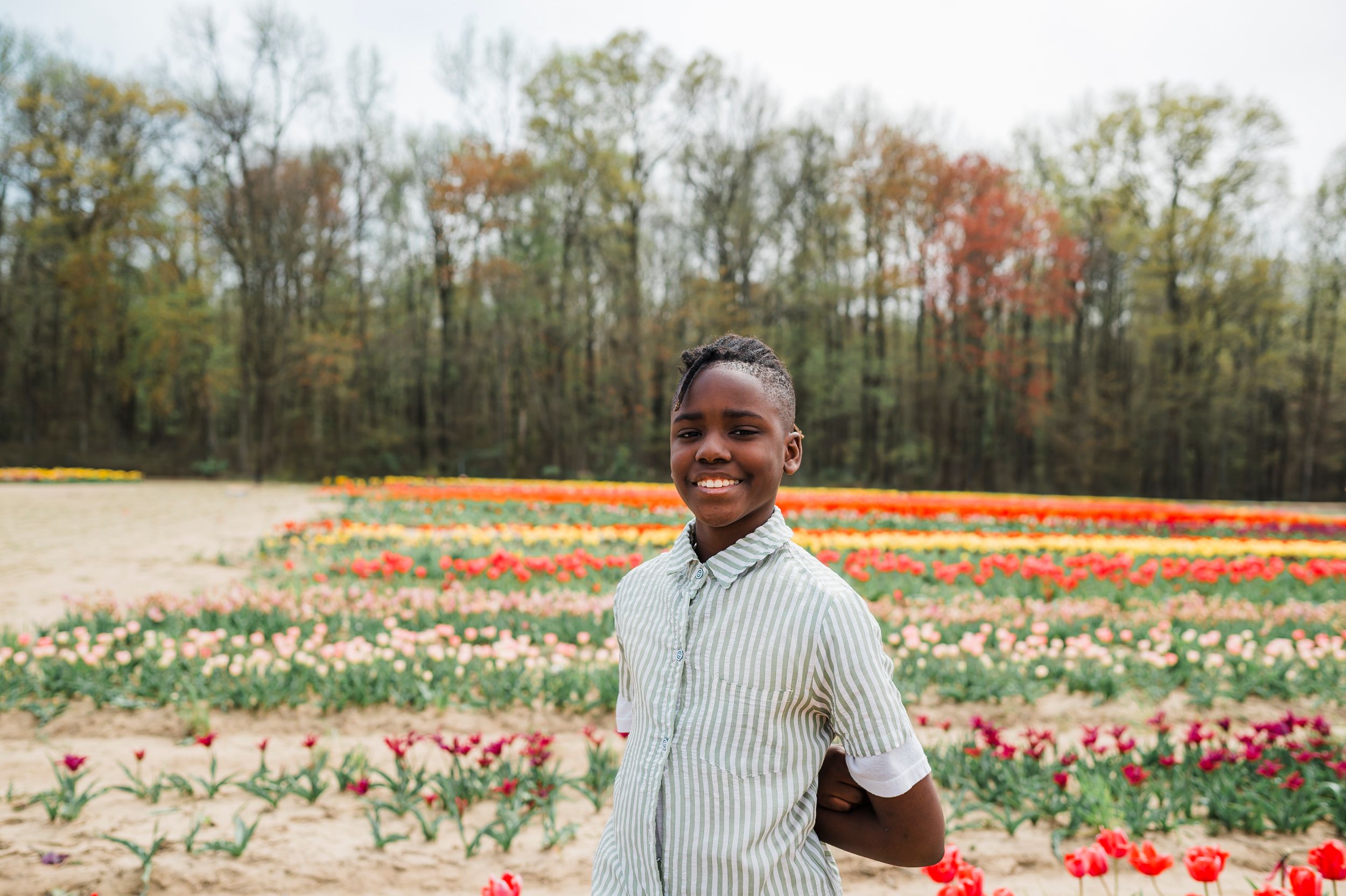 Young person in a striped shirt stands in front of colorful rows of blooming tulip flowers in a field during spring.